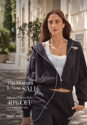 A woman in a navy tracksuit walking along a tree-lined city street while wearing wired earphones. Text: The Moment Is Now Sale. Selected Winter Styles 40% Off* Prices as marked | T&Cs Apply - Shop Now