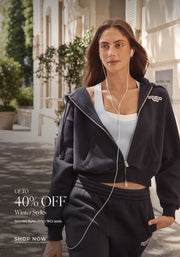 A woman wearing a navy Lorna Jane cropped zip hoodie, light blue sports tank, and matching track pants while walking outdoors with wired earphones. Text: Up To 40% Off* Winter Styles. Selected Styles Only | T&Cs apply - Shop Now
