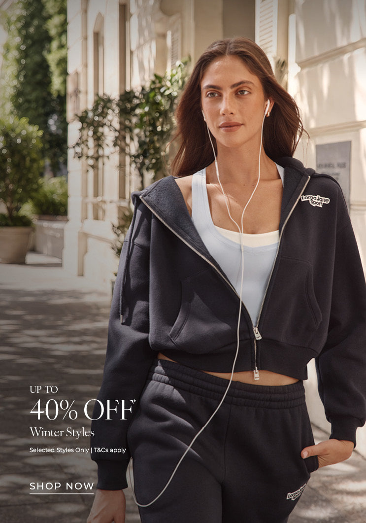 A woman wearing a navy Lorna Jane cropped zip hoodie, light blue sports tank, and matching track pants while walking outdoors with wired earphones. Text: Up To 40% Off* Winter Styles. Selected Styles Only | T&Cs apply - Shop Now