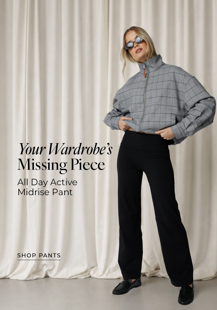 A woman in black All Day Active Midrise Pants styled with an oversized grey checked zip-up jacket, sunglasses and black loafers, against a soft neutral curtain backdrop. Text: Your Wardrobe's Missing Piece. All Day Active Midrise Pants. Shop Pants