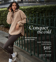 Woman wearing a beige brushed fleece half-zip pullover and dark brown thermal leggings, leaning against a green fence in an urban park. Brushed Fleece Lining. $85* T&Cs apply. Shop Now. 