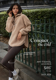 Woman wearing a beige brushed fleece half-zip pullover and dark brown thermal leggings, leaning against a green fence in an urban park. Brushed Fleece Lining. $85* T&Cs apply. Shop Now. 