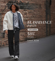 A woman walks along an urban street beside a brick wall, wearing a cream-textured jacket over a graphic T-shirt, black Flashdance pants, and sneakers. Text: Flashdance Pants Last Chance $85* T&Cs Apply. Shop Now