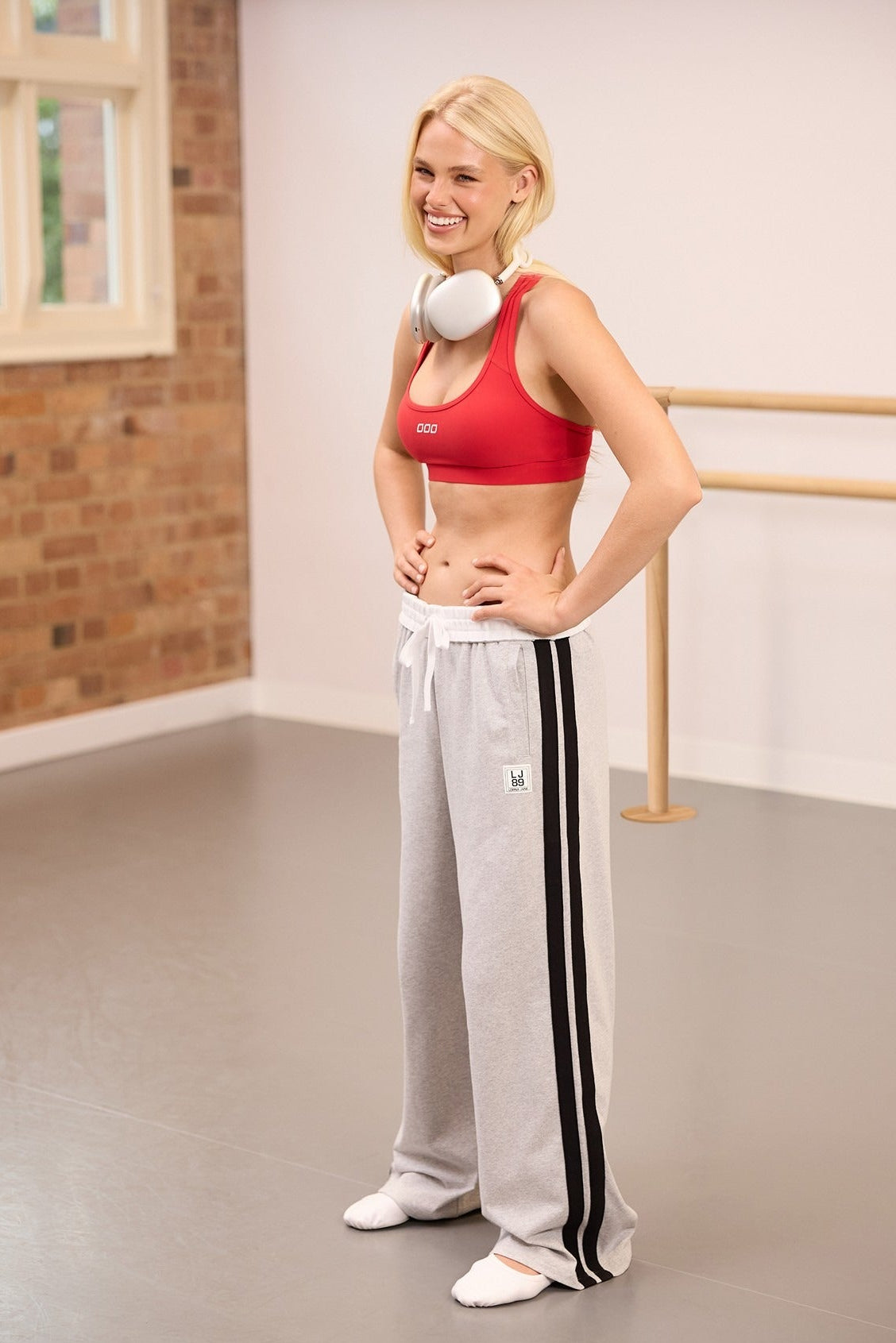Blonde woman in a red sports bra and grey striped track pants with headphones around her neck, smiling in a dance studio.