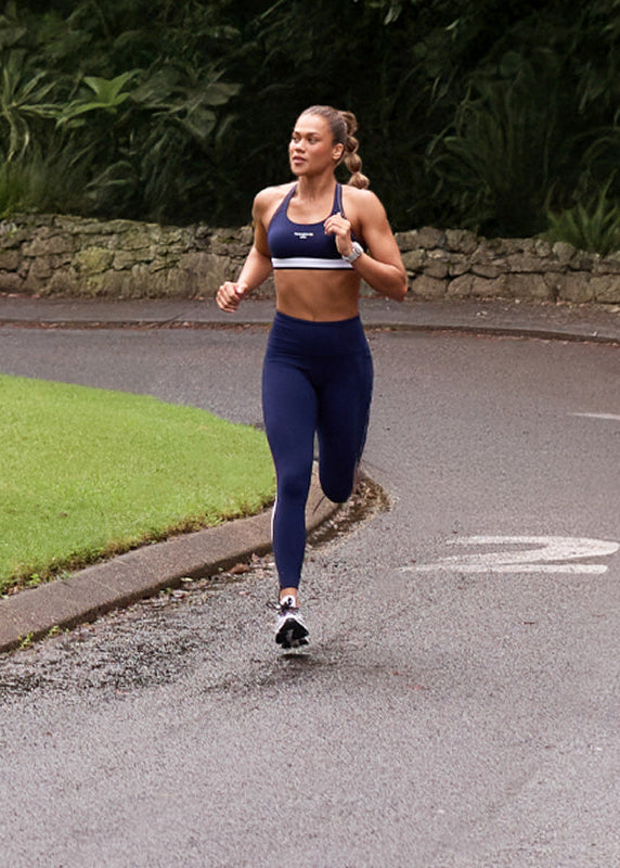 A woman jogs along a quiet, tree-lined road surrounded by lush greenery. She wears a matching blue sports bra and leggings, running through a scenic park with towering trees and a wide grassy area beside the road.