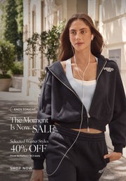 A woman wearing a navy zip-up hoodie and matching joggers over a white tank top walks outdoors while listening to wired earphones. Text: Ends Tonight. The Moment Is Now Sale. Selected Winter Styles 40% Off* Prices as marked. T&Cs Apply - Shop Now