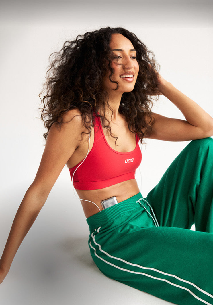 Woman wearing a red Lorna Jane sports bra and green track pants, seated against a grey studio backdrop. Text: 48 Hours Only, 15% Off* New Arrivals. T&Cs Apply - Shop Now