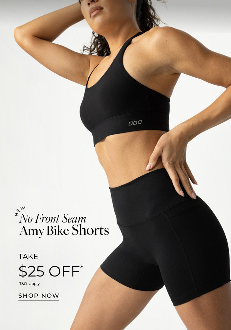 A woman posed against a plain grey studio backdrop, cropped from the chin down, wearing an all-black Lorna Jane set, a strappy sports bra and high-waisted bike shorts with her one hand on her hip, the other raised behind her head. Text: New No Front Seam Amy Bike Shorts Take $25 Off* T&Cs apply - Shop now