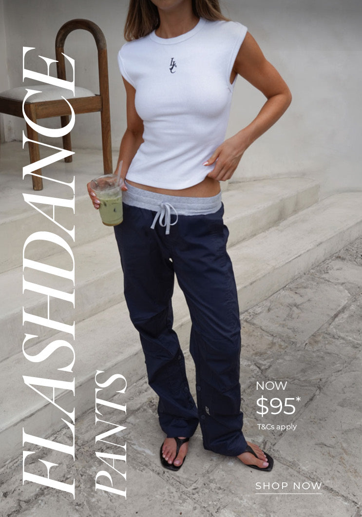 Woman wears navy Flashdance Pants with a grey drawstring waistband, styled with a white sleeveless top, sunglasses, and a navy cap, holding an iced drink in a bright, sunlit stone setting. Text: Flashdance Pants Now $95* T&Cs apply - Shop Now