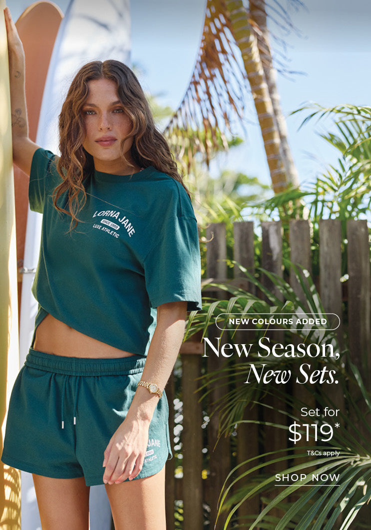 Woman in a deep teal Lorna Jane cropped tee and matching shorts, standing beside surfboards in a tropical garden. Text: New Colours Added. New Season, New Sets. Set for $119* T&Cs apply - Shop Now