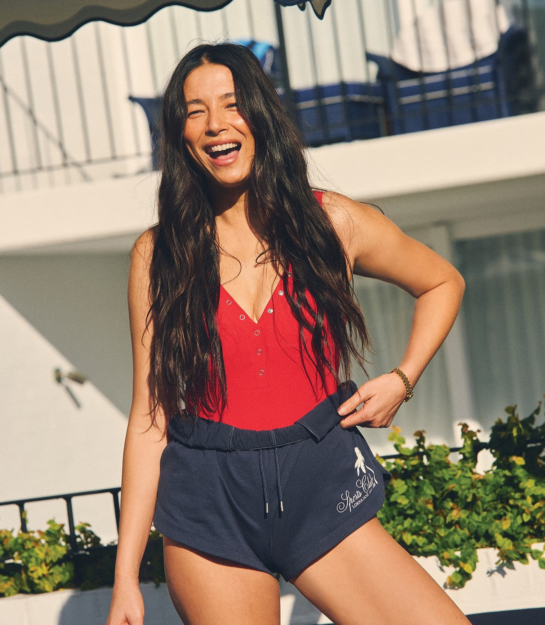 A woman laughs in the sunshine outside a poolside building, wearing a red button-front top and navy Lorna Jane Sports Club shorts.