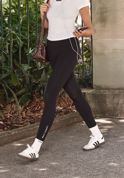 A woman walking outdoors wearing a white top, black leggings, and carrying a shoulder bag while holding a phone.