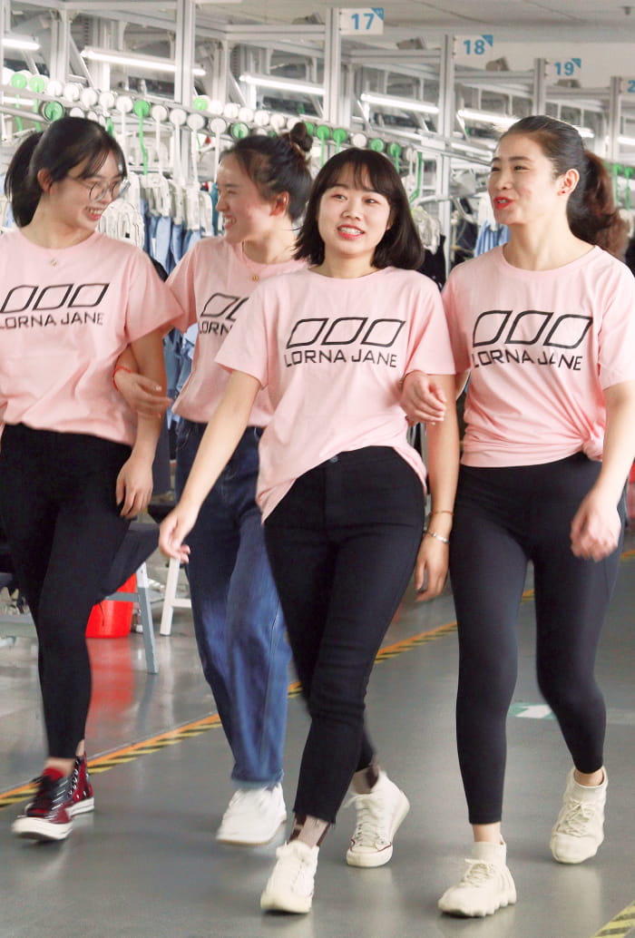 Four female garment workers in pink Lorna Jane tees walking together and smiling inside a bright manufacturing facility.