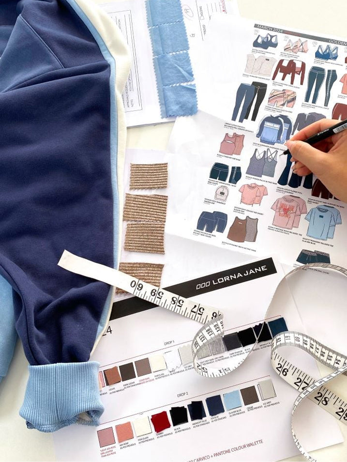 Flat lay of a Lorna Jane design workspace with a blue colour-blocked sweatshirt, fabric swatches, a garment tech sketch sheet, colour palette cards and a tape measure, as a hand sketches with a pencil.