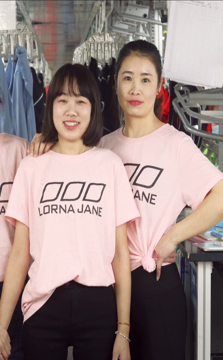 Two people wearing pink Lorna Jane T-shirts stand together in a clothing warehouse, smiling.
