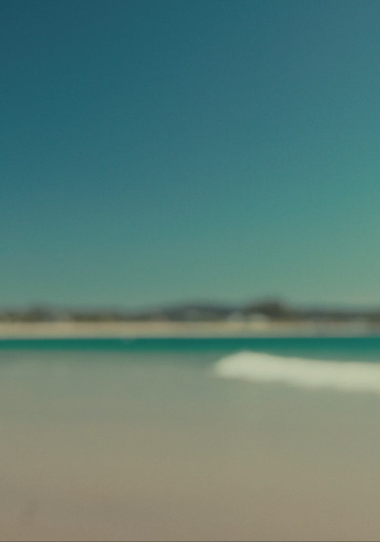 Woman wearing beachwear and activewear by the ocean on a sunny beach, with waves, rocks, and mountains in the background.