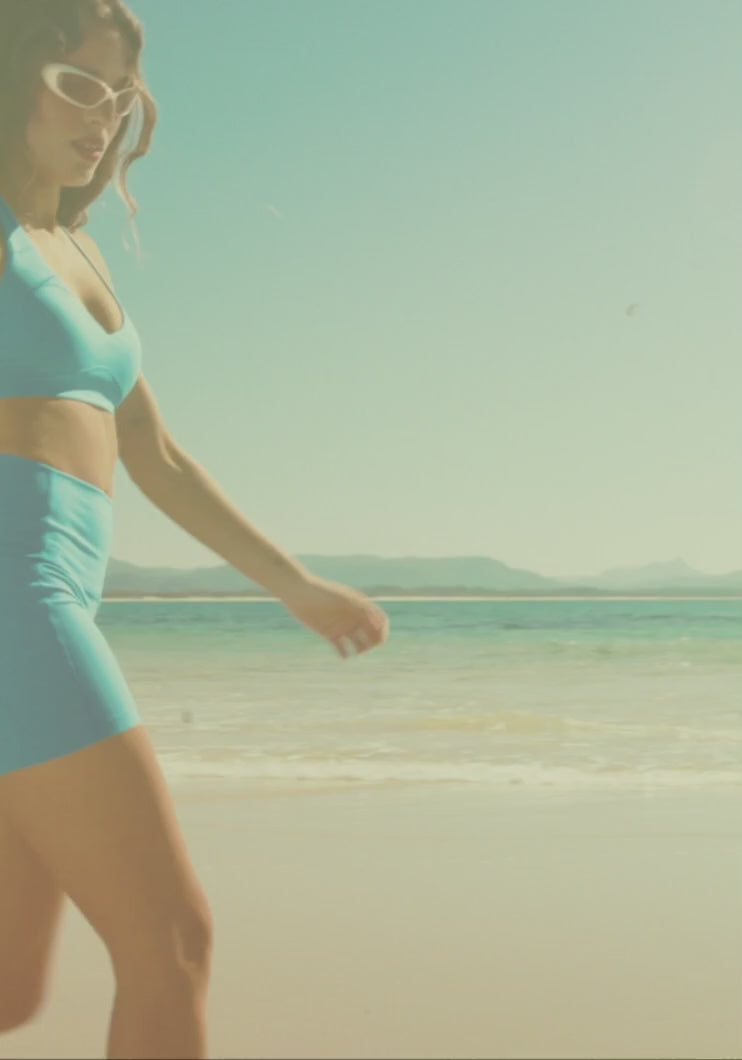 Women in colourful activewear at a sunny beach, walking along the shoreline, carrying a surfboard and playing beach volleyball.