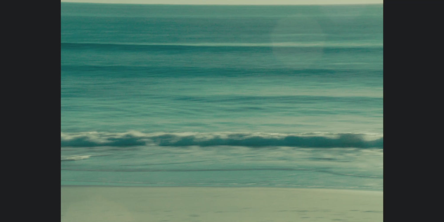 Woman wearing beachwear and activewear by the ocean on a sunny beach, with waves, rocks, and mountains in the background.