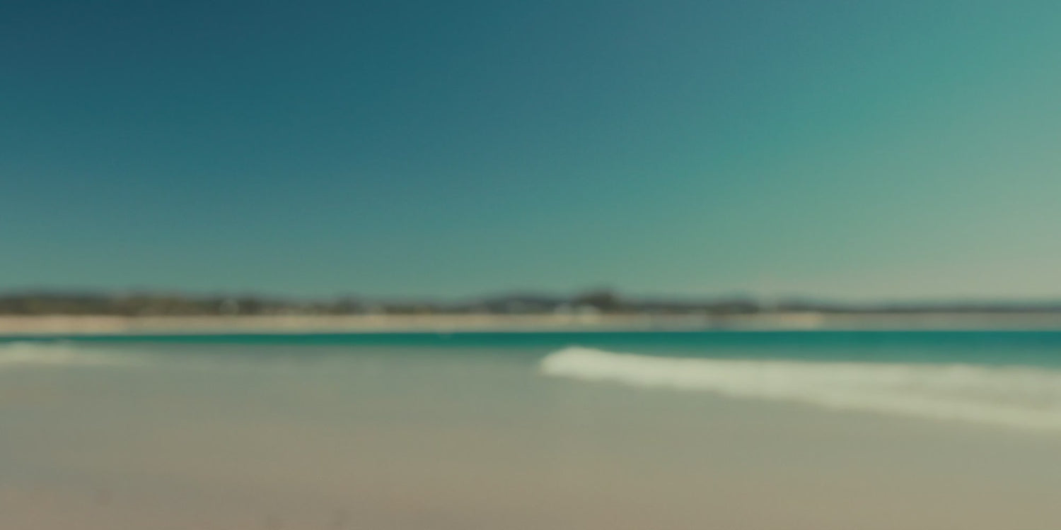 Woman wearing beachwear and activewear by the ocean on a sunny beach, with waves, rocks, and mountains in the background.