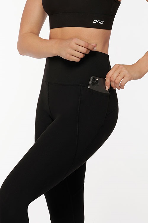 Model sliding a mobile phone into the phone pockets of black thermal leggings.