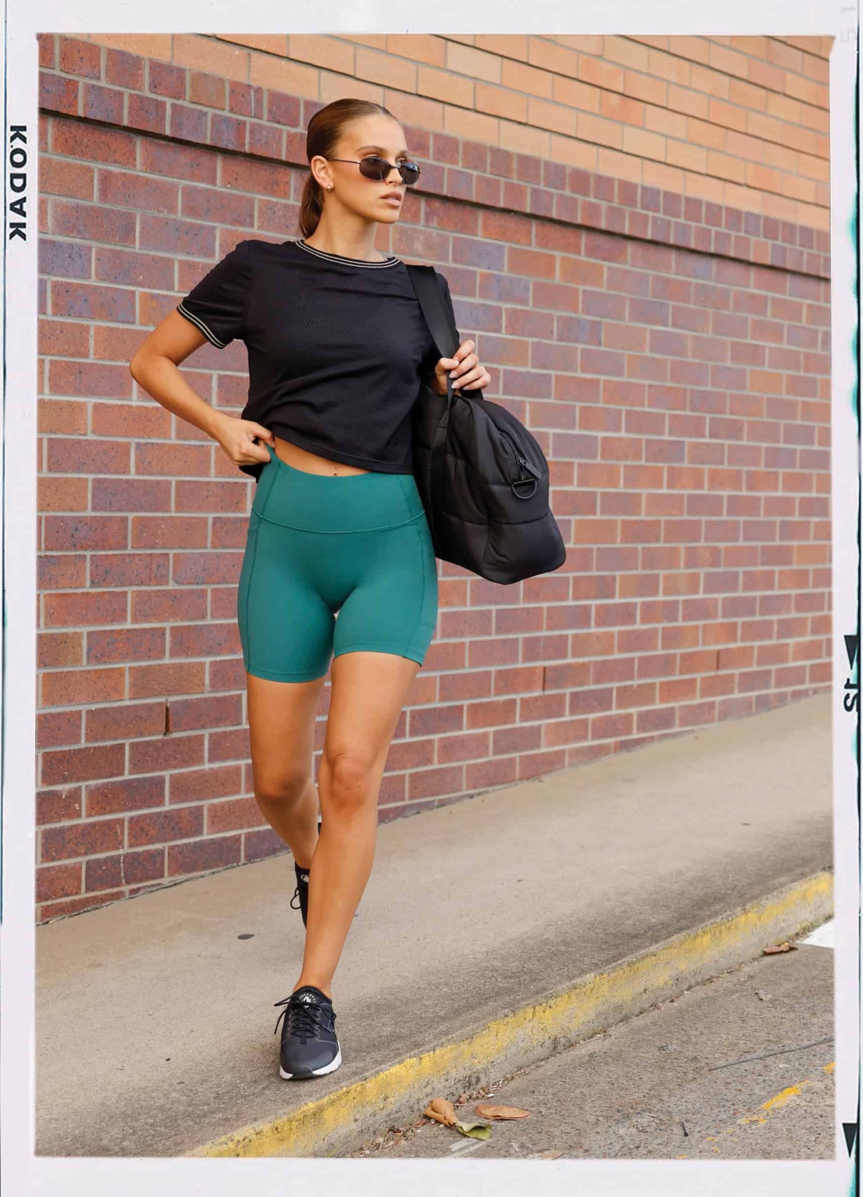 Image of model walking along the street wearing a black mesh tee with moss green bike shorts