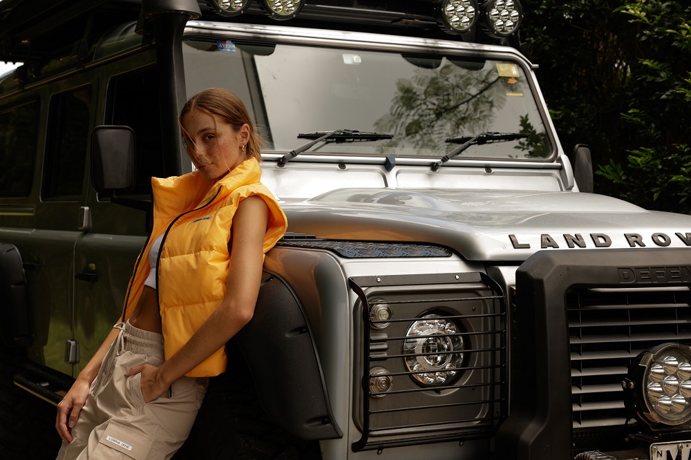 woman wearing tan cargo pants, a white sports bra and and orange puffer jacket, leaning on a truck